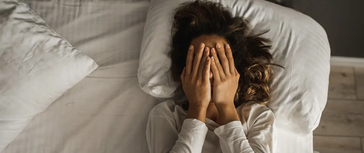Woman holding both hands over her face while laying in bed